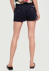 Mörkblå bomullsshorts med en figursydd design, två bakfickor och bruna knappar som accent. Stylad med en blommig topp, som visar benen och sandaler.