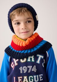 Námořnická pletená čepice a vícebarevný žebrovaný nákrčník. Nákrčník má pruhy v oranžové, červené, žluté a modré barvě. Sportovní modrý top s nápisem.