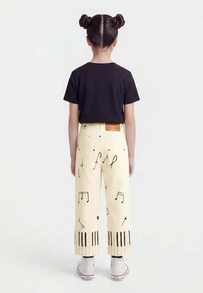 Dziecko z podwójnymi kokami, ubrane w czarny T-shirt i kremowe spodnie z nadrukiem nut i klawiszy pianina, stoi tyłem.
