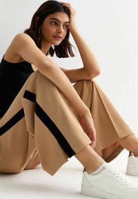 Pantaloni beige a gamba larga con bande nere laterali, realizzati in un materiale morbido, abbinati a sneakers bianche e a una canotta nera.