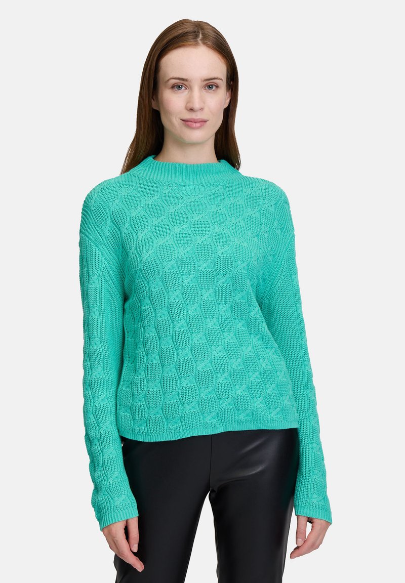 Türkiser Strickpullover mit strukturiertem Muster, rundem Ausschnitt und verlängerten Ärmeln. Kombiniert mit schwarzen, figurbetonten Hosen.