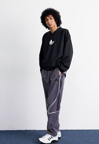 Sort hoodie med hvidt logo, grå træningsbukser med hvide striber og hvide træningssko. Sat mod en ensfarvet baggrund.