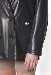 Must mustrakk, läikiv, kahekordse nööpimisega jakk, millel on tasku ja valge Adidas'i logo. Sujuv tekstuur, suured nööbid ja kohandatud lõige.