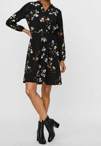 Mustassa kukkamekossa on pitkät hihat, V-mallinen pääntie, kavenettu vyötärö ja liehuva helma, jossa on valkoisia ja ruskeita kukkakuoseja.