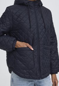 Teget prošivena jakna s patentnim zatvaračem sprijeda, kapuljačom, elastičnim manšetama i dva džepa sprijeda. Glatka tekstura i podstavljeni dizajn za toplinu.