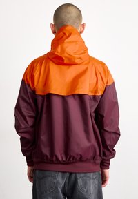Chaqueta cortavientos con una sección superior naranja y una sección inferior burdeos, con una cintura elástica y capucha para un ajuste regulable.
