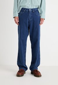 Modre jeans hlače s ravnim krojem, ki imajo sprednje žepke in gumbasto zapenje. Nosi se jih z rjavimi loafers čevlji in črtasto zeleno srajco.