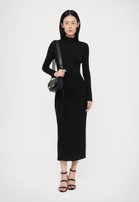 Črna obleka z dolgimi rokavi in prileganjem, z nabranimi detajli in gumbi spredaj. Usklajena z črnimi sandali na vezanje in majhno torbico.
