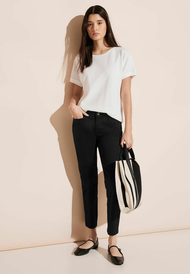 Camisetta bianca a maniche corte con texture, pantaloni neri slim-fit e ballerine nere. Borsa a tracolla a righe bianche e nere. Sfondo neutro.