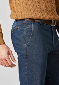 Jean en denim bleu foncé à coupe classique, avec une ceinture marron, des poches latérales et une texture subtile. Un pull en maille torsadée est visible.