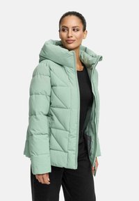 Miętowozielona kurtka puchowa z kapturem, z zamkiem z przodu, pikowanym wzorem oraz bocznymi kieszeniami. Nosi ją osoba w czarnej bluzce.