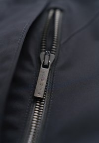 Schwarzer Stoff mit glatter Textur verfügt über einen metallischen Reißverschluss. Der Reißverschluss-Schieber hat eingravierte Details, die das elegante Design ergänzen.