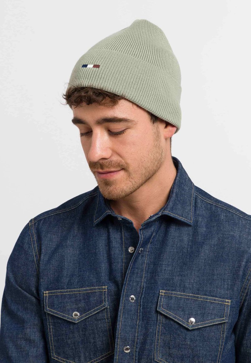 Green ribbed knit beanie with a simple design and small red, white, and blue logo detail, worn with a denim button-up shirt.
