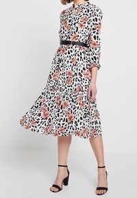 Blommig och leopardmönstrad klänning med ett svart bälte, med långa ärmar, krage med knäppning och en svävandekjol. Svarta klacksandaler.