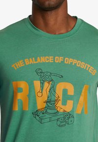 Green cotton t-shirt featuring yellow graphic text "THE BALANCE OF OPPOSITES" and an illustration of a man on a pedestal with dice.
