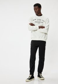 Weißer Sweatshirt mit schwarzem Text "New York City Brooklyn USA", kombiniert mit schwarzen Cargo-Hosen und schwarzen High-Top-Sneakern.