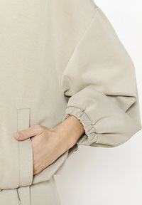 Indumento oversize di color beige chiaro con maniche voluminose e arricciate, tessuto morbido, con dettagli di tasche visibili e polsini delle maniche elasticizzati.