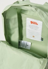 Gaiši zaļa mugursomas iekšpuse ar auduma oderi, logotipa etiķeti, kopšanas instrukcijām un birkām. Redzams apaļš šūšanas raksts.