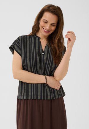 Donna con lunghi capelli castani che indossa una camicetta a righe bianche e nere a maniche corte, gonna marrone, braccialetto, sorridente con gli occhi chiusi su uno sfondo semplice.