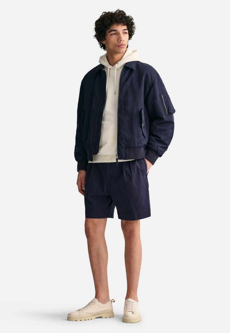 Marineblaue Bomberjacke mit lockerer Passform, kombiniert mit einem cremefarbenen Hoodie und marineblauen Shorts. Abgerundet mit beigen Sneakers, alles im minimalistischen Stil.
