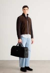 Jachetă maro din piele întoarsă cu fermoar și model reiat peste un tricou în dungi, asortată cu blugi albastru deschis și cizme negre; ținând o geantă de călătorie neagră.