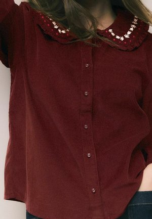 Burgundy button-up shirt with embroidered scalloped collar featuring white and red floral designs, worn with blue jeans.