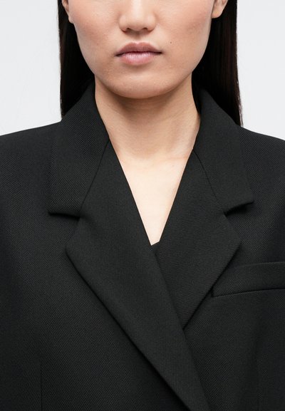 Blazer preto com um design estruturado, apresentando um colarinho com duas pontas e um único bolso no peito, feito de um tecido texturizado.