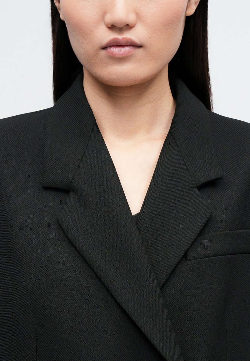 Black blazer with a structured design, featuring a notched collar and a single chest pocket, made from textured fabric.