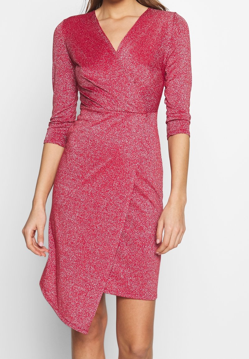 Femme portant une robe portefeuille rouge scintillante avec des manches trois-quarts et un ourlet asymétrique sur fond blanc.