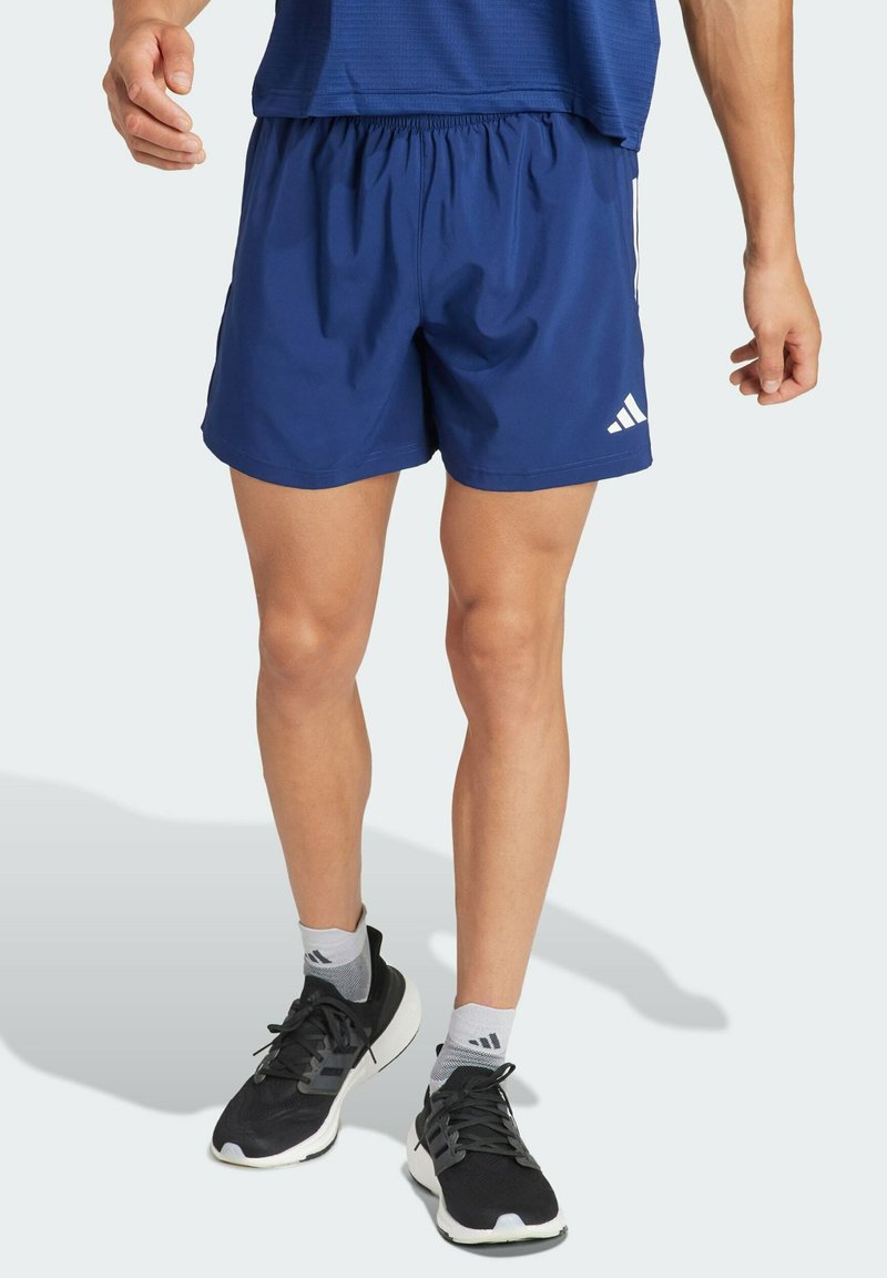 Námornícke modré športové šortky vyrobené z ľahkej látky, s elastickým pásom a tromi bielymi pruhmi po stranách. Čierne tenisky s šedými ponožkami.