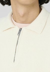 Pull en zip dans un blanc cassé, avec une texture douce, un col plat et une fermeture éclair argentée avec un logo. Superposé sur une chemise rose.