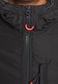 Giacca nera con colletto alto e chiusura con zip, caratterizzata da un accento rosso e un cordino regolabile per una vestibilità aderente. Tessuto strutturato.