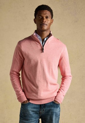 Junger Mann trägt einen hellrosa Pullover mit Reißverschluss bis zur Brust über einem blau gestreiften Hemd, steht mit den Händen in den Taschen vor einem einfarbigen beigen Hintergrund.