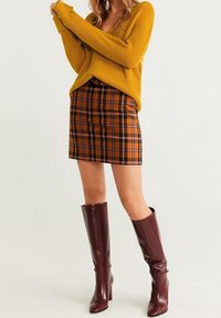 Frau in senfgelbem Pullover, braun-schwarzem kariertem Minirock und knielangen, dunkelbraunen Lederstiefeln, die vor einem schlichten Hintergrund steht.