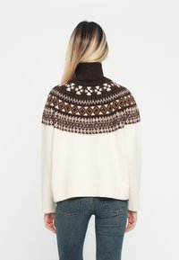 Sweater creme com um peito texturizado em padrão castanho, apresentando um colarinho alto e mangas longas, combinado com jeans skinny.