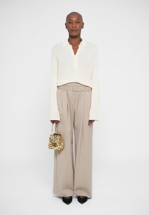 Woman in beige wide-leg pants with belt, white ribbed knit sweater, black pointed shoes, holding small gold sequin handbag, against white background.