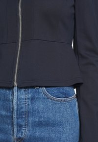 Granatowa kurtka na zamek błyskawiczny o fasonie dopasowanym w talii i rozszerzającym się ku dołowi; gładka tekstura i długie rękawy, połączona z niebieskimi dżinsami z kontrastowym przeszyciem.