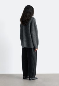 Grauer, flauschiger Pullover mit lockerer Passform, kombiniert mit weiten schwarzen Hosen und schwarzen Schuhen, von hinten betrachtet. Minimalistischer Hintergrund.