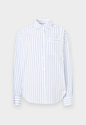 Vrouw staat en draagt een wit met blauw verticaal gestreept shirt met lange mouwen en bijpassende losse shorts, met zwarte platte schoenen met witte stippen.