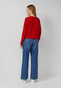 Roter gerippter Pullover mit überschnittenen Schultern, kombiniert mit weit geschnittenen blauen Jeans. Das Outfit wird mit weißen Sneakern abgerundet.