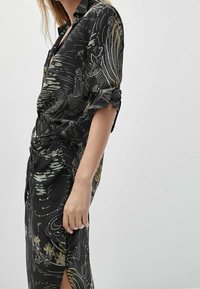 Robe noire avec des motifs floraux et de vagues dorées. Manches longues retroussées, design à col, fente sur le côté et tissu lisse.