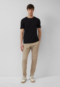 Schwarzes T-Shirt mit kurzen Ärmeln, beiges Slim-Fit-Hosen und beigefarbene Sneakers mit strukturierten Akzenten. Das Model steht aufrecht vor einem neutralen Hintergrund.