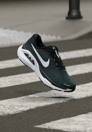 Zwart-groene sportschoen met mesh bovenzijde, witte accenten, gestructureerde zool en zichtbare "ZoomX"-branding, geplaatst boven een zebrapad.