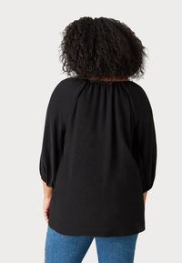 Top leggero e nero con scollo arricciato, maniche a tre quarti ampie e vestibilità rilassata. Indossato con jeans in denim blu.