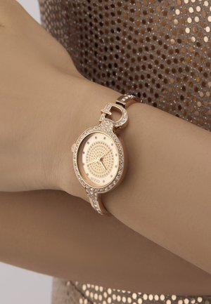 Rožinio aukso laikrodis su apvalia dėže, puoštu skaidriais kristalais ir dekoratyvia juosta su užsegimu. Ciferblatas turi blizgių detalių.