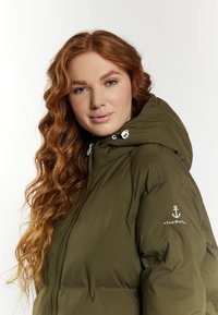 Jachetă puffer verde măsliniu cu glugă, cu închidere prin fermoar. Include un accent cu logo pe manșetă și o țesătură texturată pentru căldură.