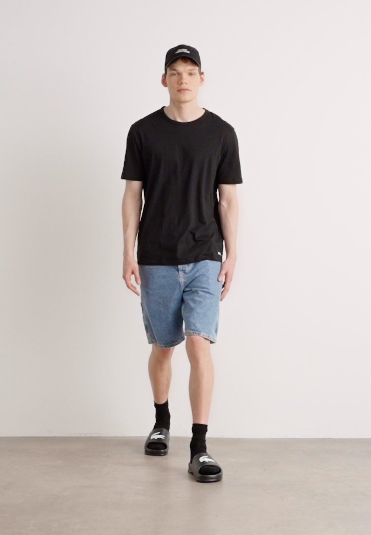 T-shirt noir, short en denim, chaussettes noires et sandales à enfiler. Le mannequin se tient sur un sol en béton clair avec un mur blanc uni en arrière-plan.