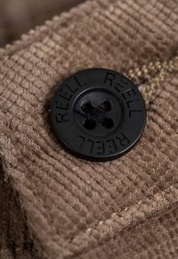 Schwarzer Vierlochknopf mit dem eingeprägten Schriftzug "REELL", befestigt an einem strukturierten braunen Kordstoff, der ein dezentes Streifenmuster aufweist.