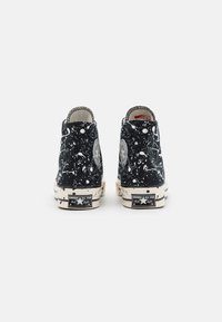 Tênis pretos de cano alto com um padrão branco salpicado, solas de borracha e a marca Converse no calcanhar. O design apresenta um patch de logotipo redondo.