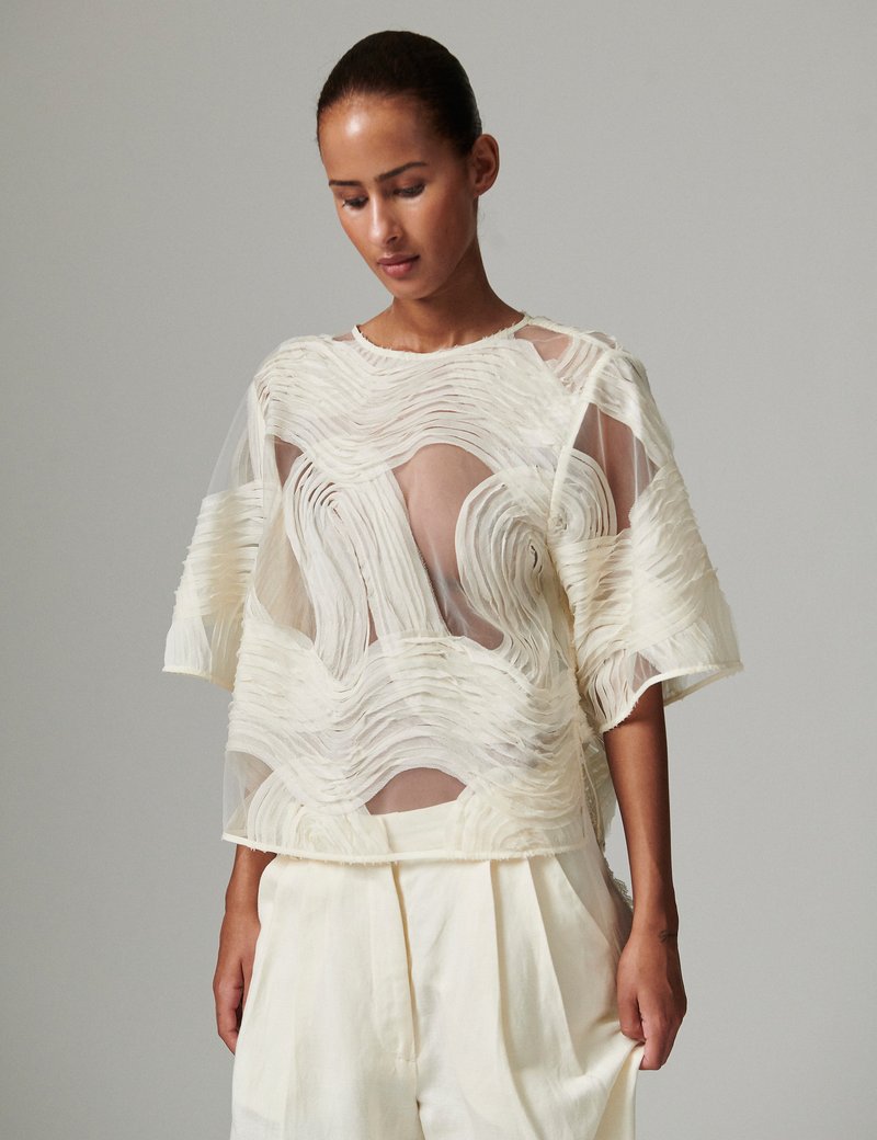 Κρεμ διάφανο τοπ με υφασμάτινες λεπτομέρειες σε κυματιστή υφ ή. Χαλαρή εφαρμογή, κοντά μανίκια, φορεμένο πάνω από ανοιχτόχρωμο κάτω μέρος. Απαλό, ρέον υλικό.